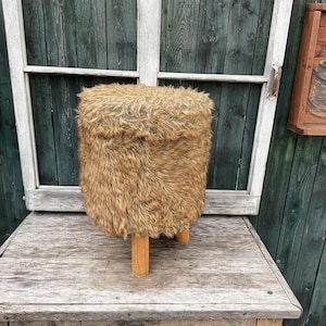 Pu&ograve; includere: Uno sgabello peloso marrone e beige con quattro gambe in legno. Lo sgabello &egrave; su una superficie in legno.