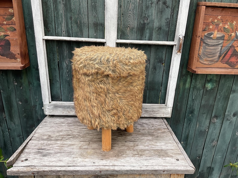Pu&ograve; includere: Uno sgabello peloso marrone e beige con quattro gambe in legno. Lo sgabello &egrave; su una superficie in legno.