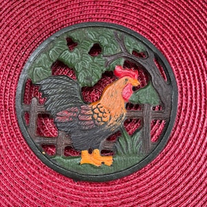 May include: A circular cast iron trivet with a detailed rooster design. The rooster is depicted in shades of orange, black, and red, set within a green landscape with a fence and tree. The trivet is placed on a red, textured mat.
