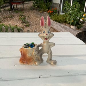 Może przedstawiać: Ceramiczna figurka Bugs Bunny'a stojąca obok brązowego pnia drzewa z kolekcja kolorowych ołówków w środku. Figurka jest biała z różowymi uszami i jasnoszarym ciałem.