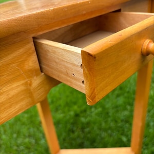 May include: A light brown wooden console table with a smooth, polished finish. The table features a single rectangular drawer with a small, round wooden knob handle. The drawer is partially pulled open, revealing the interior construction.