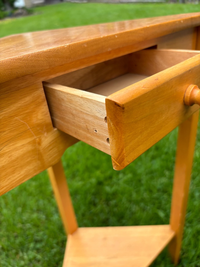May include: A light brown wooden console table with a smooth, polished finish. The table features a single rectangular drawer with a small, round wooden knob handle. The drawer is partially pulled open, revealing the interior construction.