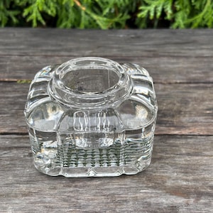 May include: A clear, square glass inkwell with a circular opening. The inkwell has a decorative, textured design and sits on a wooden surface. The background features green foliage.
