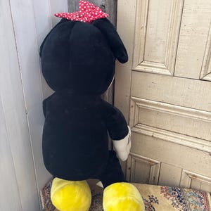May include: A large plush toy of a cartoon character, featuring a black body, yellow feet, white gloves, and a red bow with white polka dots. The toy is seated on a patterned rug.