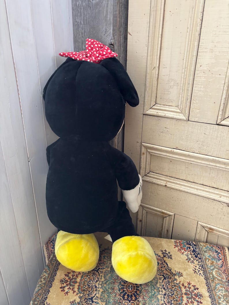 May include: A large plush toy of a cartoon character, featuring a black body, yellow feet, white gloves, and a red bow with white polka dots. The toy is seated on a patterned rug.