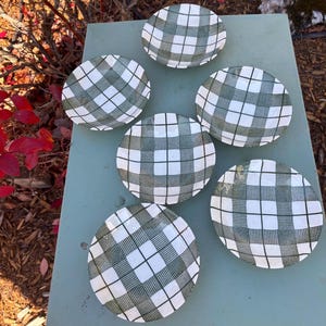 May include: Six round ceramic plates with a green and white chequered pattern. The plates are arranged on a light green surface, with red leaves and brown branches in the background. The plates are a classic design.