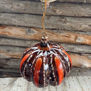 Könnte beinhalten: Eine braune und orangefarbene Keramik-Pendelleuchte in Form eines Kürbisses mit Löchern über die gesamte Oberfläche. Die Leuchte hängt an einer goldenen Kette.