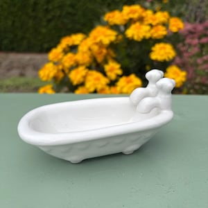 以下が含まれることがあります： 猫足バスタブの形をした白い陶器製のソープディッシュ。滑らかで光沢のある仕上げで、片端に2つの彫刻された蛇口ハンドルがあり、底面には小さな丸い脚が付いています。石鹸などの小物を置くためのデザインです。
