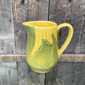 May include: A yellow ceramic jug with a green corn design. The jug has a handle and a wide mouth.