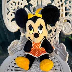 K&ouml;nnte beinhalten: Eine Minnie Mouse Pl&uuml;schpuppe mit schwarzem Fell, gro&szlig;en schwarzen Ohren und einer gelben Schleife. Sie tr&auml;gt ein orangefarbenes Kleid mit wei&szlig;en Punkten und gelbe Schuhe. Die Puppe sitzt auf einem wei&szlig;en, verzierten Metallstuhl.