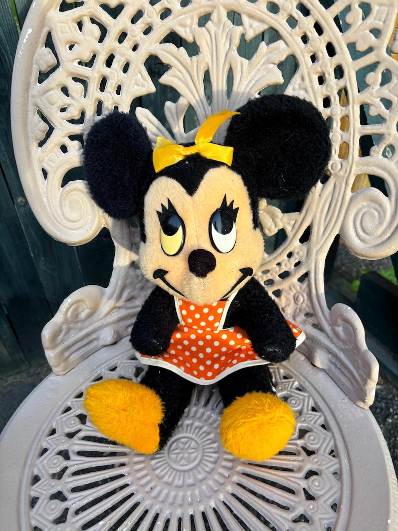 K&ouml;nnte beinhalten: Eine Minnie Mouse Pl&uuml;schpuppe mit schwarzem Fell, gro&szlig;en schwarzen Ohren und einer gelben Schleife. Sie tr&auml;gt ein orangefarbenes Kleid mit wei&szlig;en Punkten und gelbe Schuhe. Die Puppe sitzt auf einem wei&szlig;en, verzierten Metallstuhl.