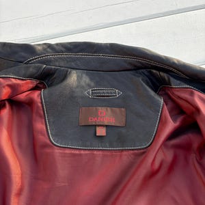 May include: The inside of a black leather jacket with a red lining. A brown leather tag with white stitching reads "D.  DANIER".