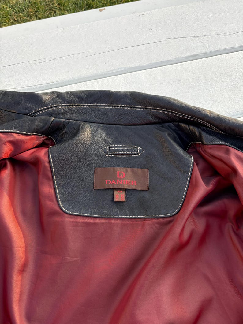 May include: The inside of a black leather jacket with a red lining. A brown leather tag with white stitching reads "D.  DANIER".