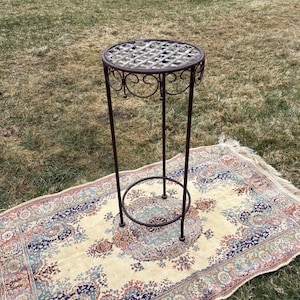 Peut inclure: Un support de plante en fer forgé, de couleur marron foncé, avec un dessus circulaire à motif de treillis. Le support présente des volutes décoratives. Il mesure environ 76 cm de haut et est posé sur un tapis à motifs.