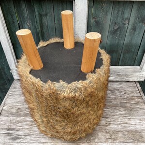Pu&ograve; includere: Uno sgabello peloso marrone con tre gambe in legno. Lo sgabello &egrave; su una superficie in legno.