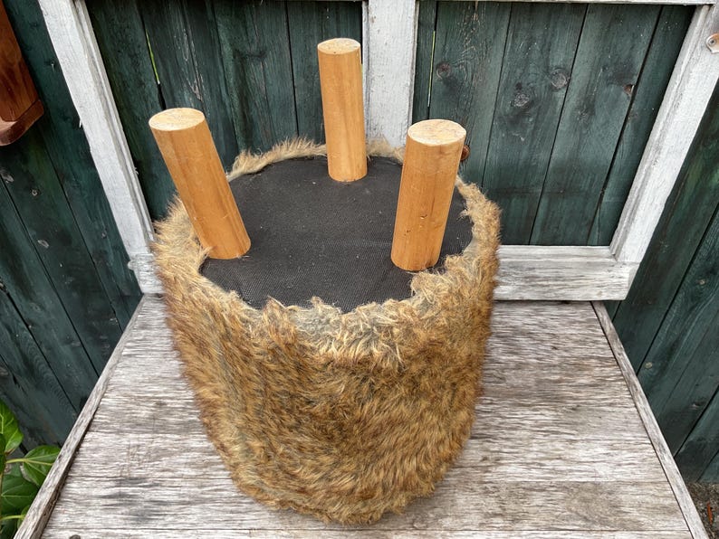 Pu&ograve; includere: Uno sgabello peloso marrone con tre gambe in legno. Lo sgabello &egrave; su una superficie in legno.