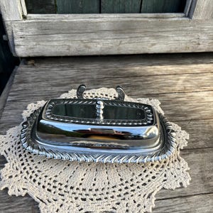 Puede incluir: Un mantequillero de plata con tapa de vidrio y borde decorativo. El plato está sobre un encaje de ganchillo.