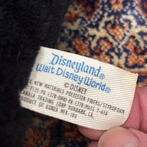 May include: A close-up of a tag from a Disneyland Walt Disney World plush toy. The tag is white with blue text that reads "Disneyland Walt Disney World" and "&copy; DISNEY". The tag also includes material and manufacturing information.