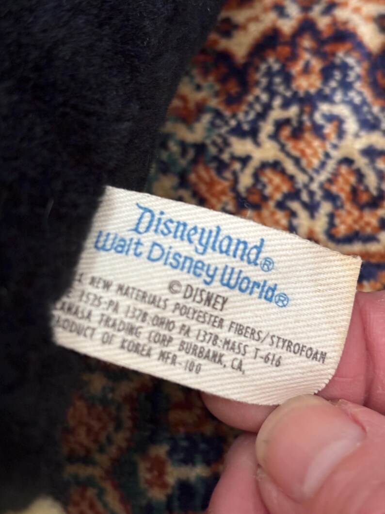 May include: A close-up of a tag from a Disneyland Walt Disney World plush toy. The tag is white with blue text that reads "Disneyland Walt Disney World" and "&copy; DISNEY". The tag also includes material and manufacturing information.