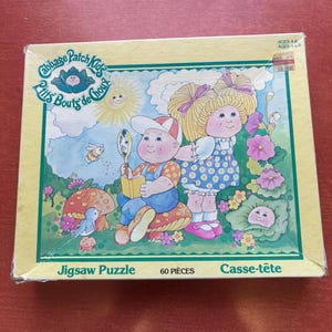 May include: A Cabbage Patch Kids jigsaw puzzle box. The box features a colourful illustration of two Cabbage Patch Kids in a whimsical garden setting with flowers, mushrooms, and a smiling sun. The puzzle has 60 pieces.