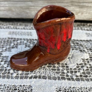 May include: A small, ceramic cowboy boot with a glossy finish. The boot is primarily brown with red accents on the front. The boot is a decorative item, likely a small vase or container.