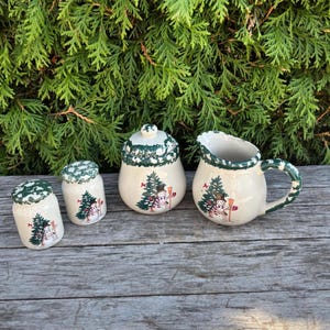 May include: A cream-colored ceramic kitchen set with a winter theme. The set includes a sugar bowl, creamer, and two small jars, all decorated with green pine trees and snowmen. The rims and lids have a green, textured design.