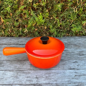 May include: A bright orange enamelled cast iron saucepan with a matching lid and a single long handle. The pot features a smooth, glossy finish and a small, black circular knob centred on the lid. The design is round with a deep basin.