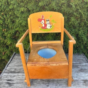 May include: A vintage wooden potty chair with a light brown finish. The chair features a blue basin and a backrest decorated with a cartoon image of a deer and rabbits. The chair has armrests and is set on a wooden surface.