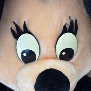 May include: Close-up of a plush toy face with large, oval eyes and black eyelashes. The toy has a peach-colored face, a black nose, and a red and white polka-dot bow. The toy is a cartoon character.