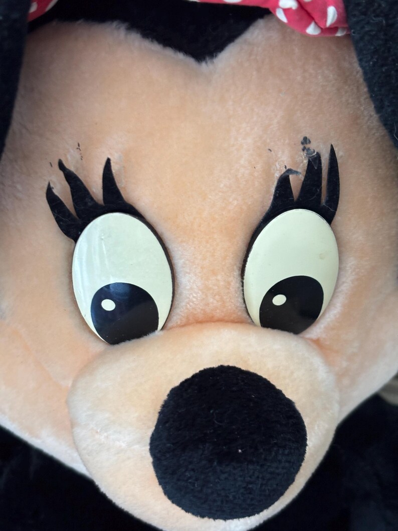 May include: Close-up of a plush toy face with large, oval eyes and black eyelashes. The toy has a peach-colored face, a black nose, and a red and white polka-dot bow. The toy is a cartoon character.