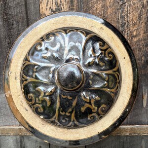Puede incluir: Una tapa de cerámica circular con un esmalte negro brillante y oscuro con patrones de volutas en relieve de color tostado. El centro tiene un pomo liso y elevado, y el borde exterior es una banda ancha y plana de arcilla beige.