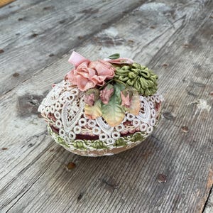 Puede incluir: Un adorno decorativo con forma de huevo cubierto de encaje blanco y adornado con flores de tela rosa y verde.