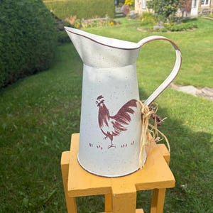 Peut inclure: Un pichet en métal de couleur crème avec une finition mouchetée et une illustration de coq marron sur le devant. Le pichet présente un large bec verseur, une anse incurvée et un bouquet décoratif en raphia attaché à la base de l'anse.
