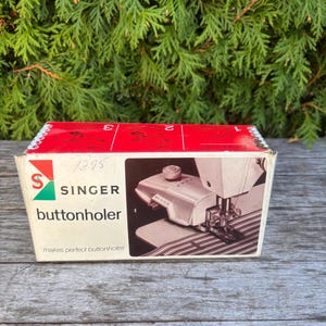 Può includere: Una scatola per asolatrice Singer vintage con la parte superiore rossa e i lati bianchi. La scatola presenta il logo Singer e le parole "buttonholer" e "makes perfect buttonholes". La scatola è posta su uno sfondo di fogliame verde.