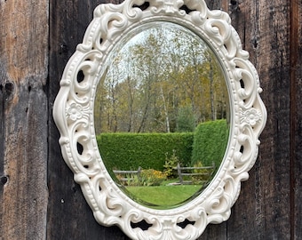 May include: An oval, off-white ornate mirror with a decorative frame. The mirror reflects a green hedge, trees, and a wooden fence. The mirror is mounted on a weathered, dark brown wooden wall.