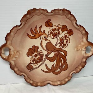 May include: A decorative ceramic serving tray with a scalloped edge and two handles. The tray features a brown and white design of a bird with a long tail and floral accents.