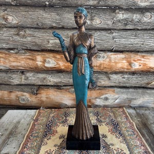 May include: A bronze art deco sculpture of a woman in a teal dress and matching gloves, posed with one arm extended. The sculpture has a bronze-colored shawl and a head wrap. It stands on a black base, with a patterned rug in the foreground.