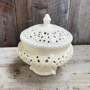 May include: Cream ceramic lidded jar with an elaborate openwork design. The jar has a round base, a rounded body, and a domed lid with a small finial. The openwork pattern gives a delicate, airy look, suitable for decorative storage.