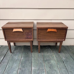 Puede incluir: Un par de mesitas de noche de estilo moderno de mediados de siglo. Cada mesita está hecha de madera marrón oscuro con un cajón y un tirador curvo de madera clara. Las mesitas tienen patas cónicas y forma rectangular. Una mesita muestra desgaste en el cajón.