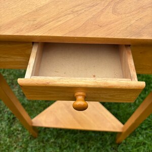 May include: A light brown wooden corner table featuring a triangular top, a single pull-out drawer with a small round knob, and a lower triangular shelf. The table is constructed from wood with a visible grain pattern and a smooth finish.