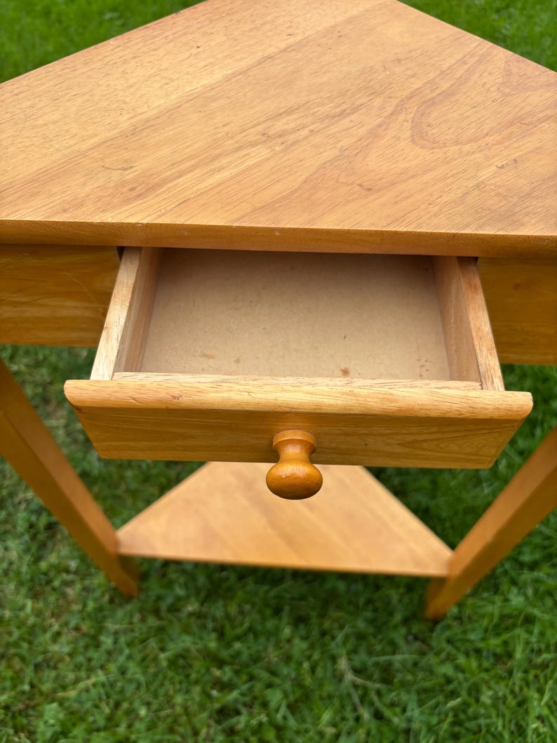 May include: A light brown wooden corner table featuring a triangular top, a single pull-out drawer with a small round knob, and a lower triangular shelf. The table is constructed from wood with a visible grain pattern and a smooth finish.