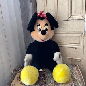 May include: A plush Minnie Mouse doll with a black body, white gloves, and a red polka dot bow. The doll has yellow spheres for feet and is seated on a patterned rug.