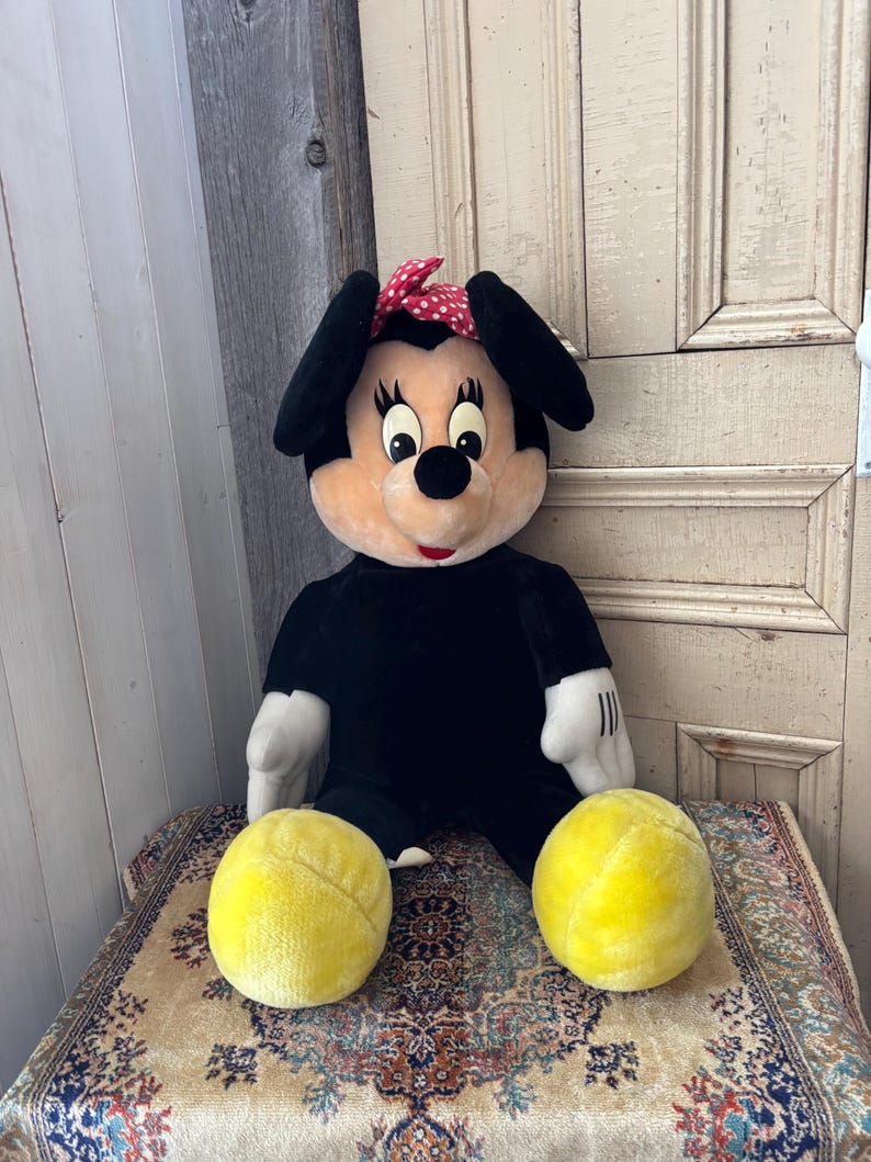 May include: A plush Minnie Mouse doll with a black body, white gloves, and a red polka dot bow. The doll has yellow spheres for feet and is seated on a patterned rug.
