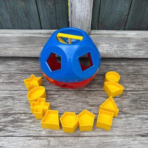 Puede incluir: Un juguete clasificador de formas colorido. El juguete es una esfera azul y roja con agujeros de varias formas. Formas amarillas, incluyendo estrellas, círculos y cuadrados, están dispuestas alrededor de la esfera. Una manija amarilla está en la parte superior.