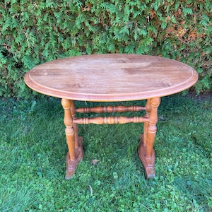 May include: An oval wooden side table with a smooth, light brown surface. The table features four turned legs connected by two horizontal support bars, resting on two flat, wide feet that provide stability to the overall structure.