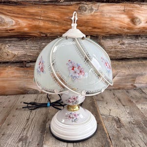 Peut inclure: Une lampe de table vintage avec une base blanche et un motif floral. La lampe a un abat-jour en verre avec un motif floral rose et rouge. Elle a un cadre en métal blanc et un cordon d'alimentation noir.
