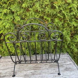 Pode incluir: Revisteiro em ferro forjado preto com um design decorativo de rolagem. O revisteiro tem uma forma retangular com barras verticais e pernas curvas. O fundo é uma sebe verde.