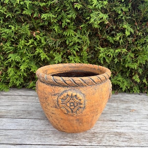 May include: A round, terracotta-coloured planter with a weathered, rustic finish. The rim features a raised, twisted rope-like texture, and the exterior is decorated with a central, circular medallion containing a symmetrical floral relief pattern.