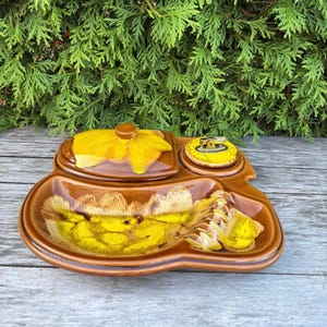 Puede incluir: Un cenicero vintage de cerámica marrón con un diseño floral amarillo. El cenicero tiene una tapa, un soporte para encendedor y un plato. El cenicero está sobre una superficie de madera con follaje verde en el fondo.
