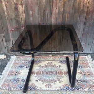 May include: A square side table featuring a dark smoked glass top and a continuous black tubular metal frame. The base consists of a single curved metal structure that supports the glass surface, creating a cantilevered effect with a sleek finish.